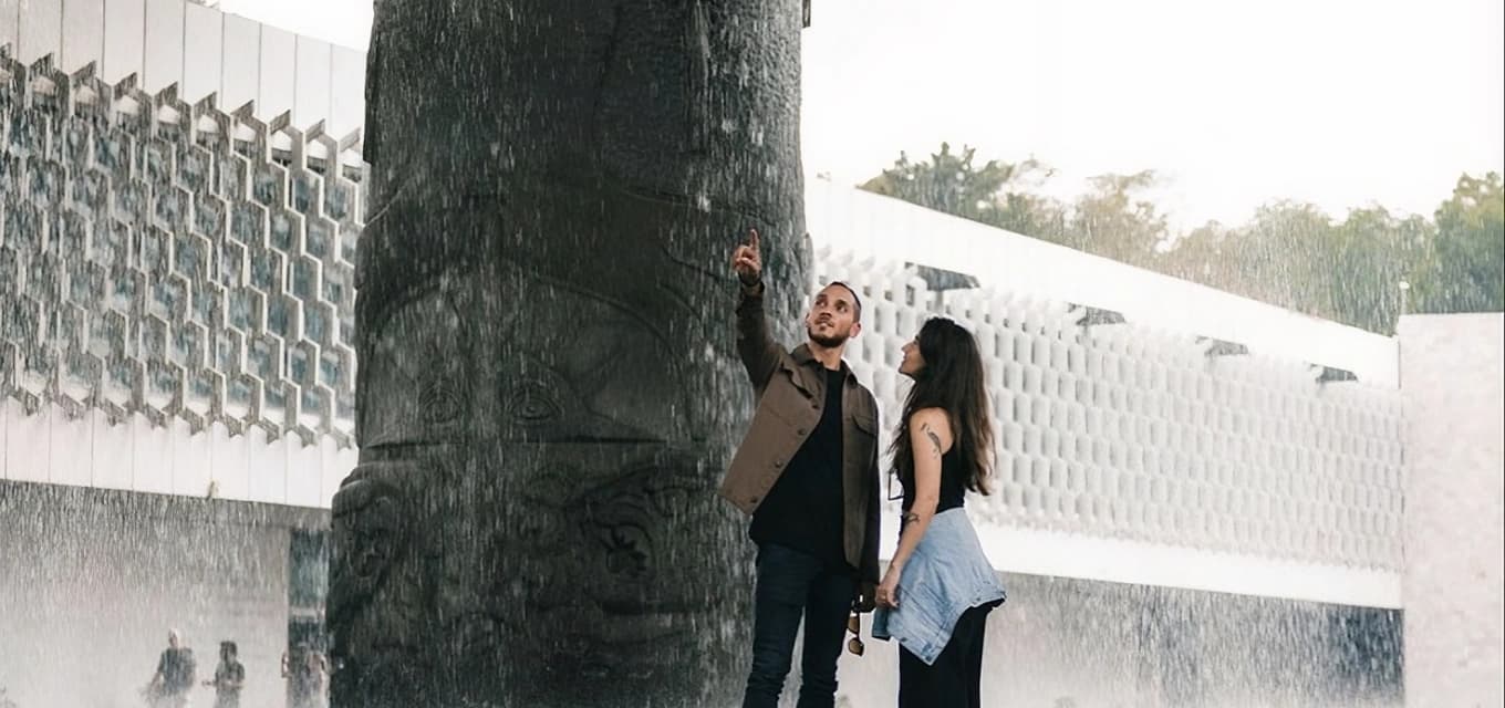 A couple under the "El Paraguas" rain, reaching to touch the water falling from the carved column at the National Museum of Anthropology