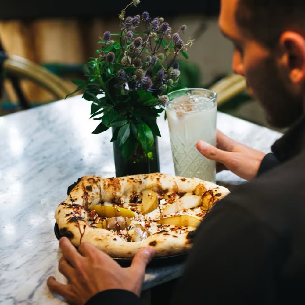 A diner before a pear pizza on Aura's marble table, beside a cut-glass tumbler with ice and a vase of blue thistles