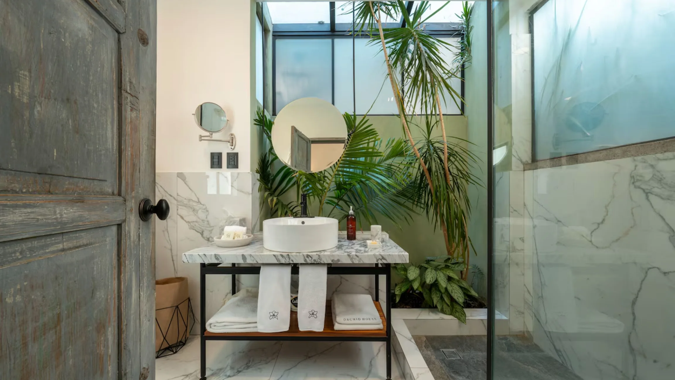 A bathroom with a vessel basin on marble, a round mirror and a palm beneath the skylight