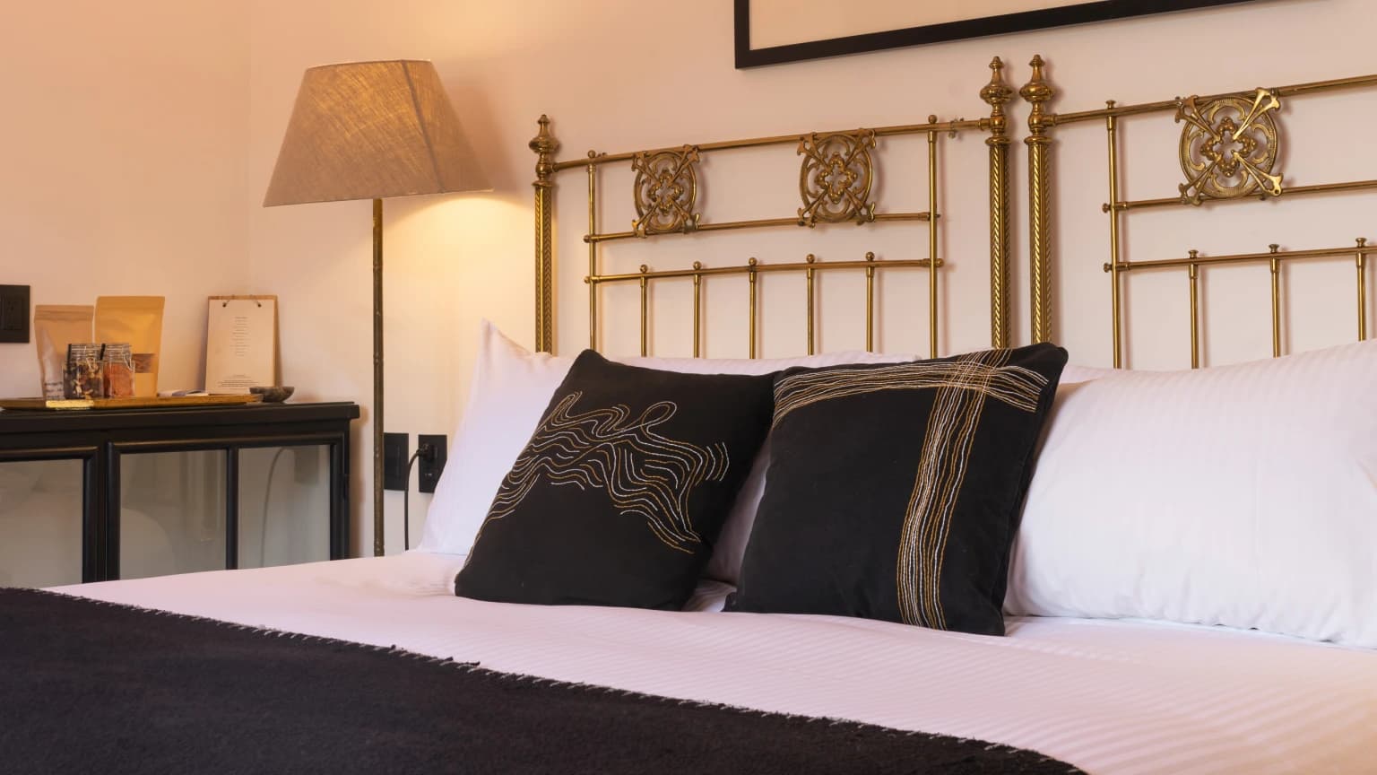A carved brass headboard with embroidered cushions and a floor lamp beside the bed