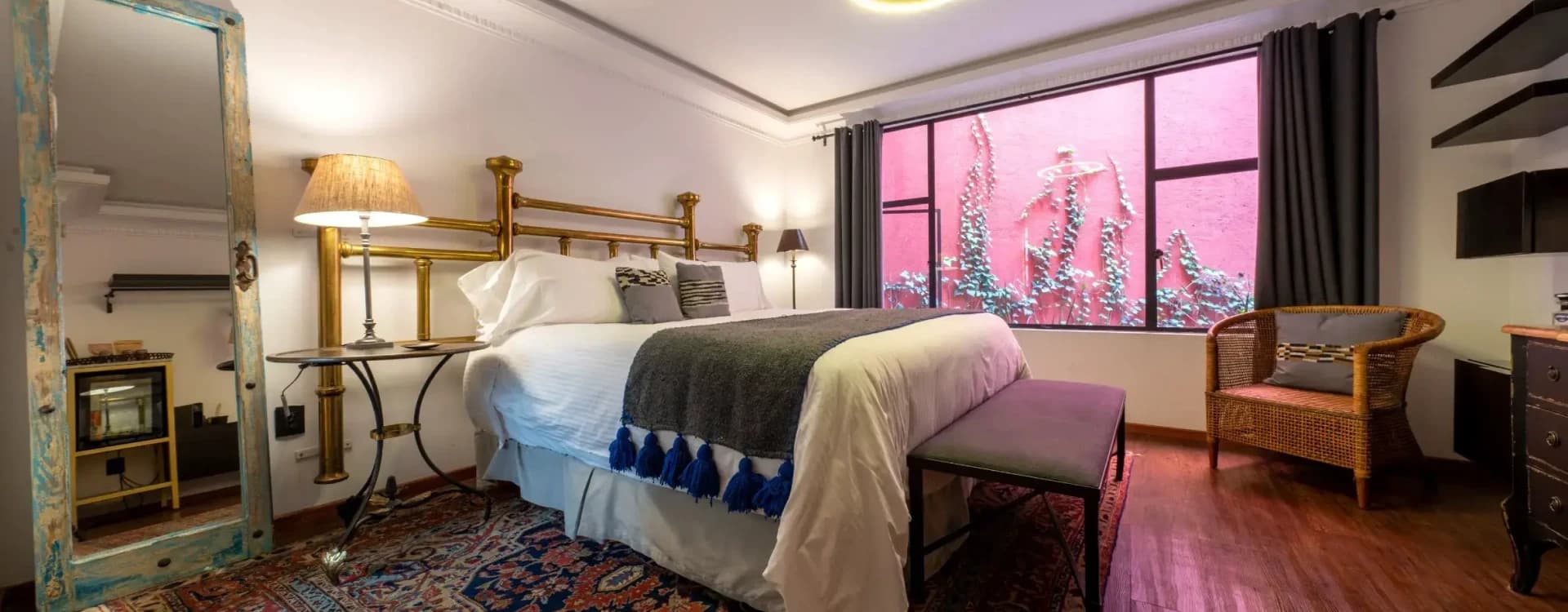 Bed with a brass headboard, a Persian rug and a window onto a pink wall with climbing vines