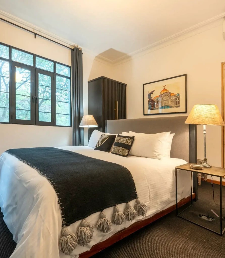 King bed with a grey headboard, a framed print of the Palacio de Bellas Artes and a black wardrobe beside the window