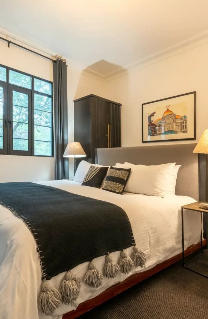 King bed with a grey headboard, a framed print of the Palacio de Bellas Artes and a black wardrobe beside the window
