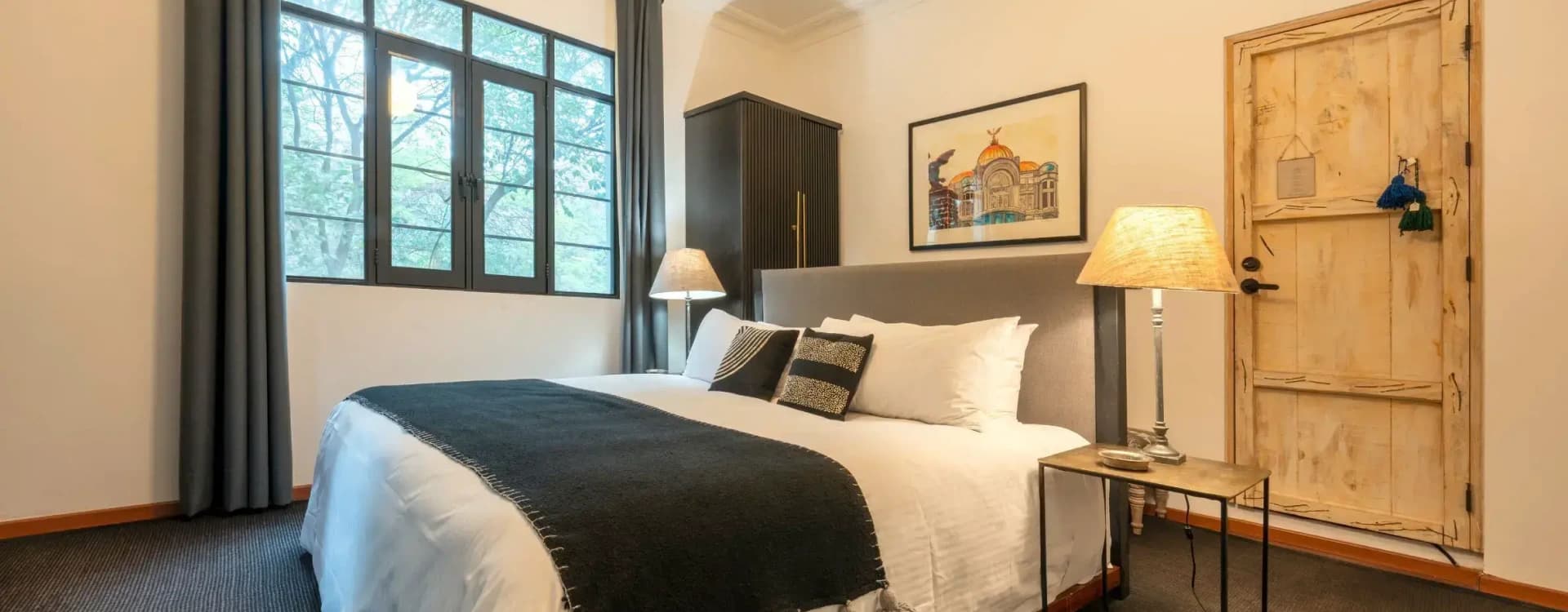 King bed with a grey headboard, a framed print of the Palacio de Bellas Artes and a black wardrobe beside the window