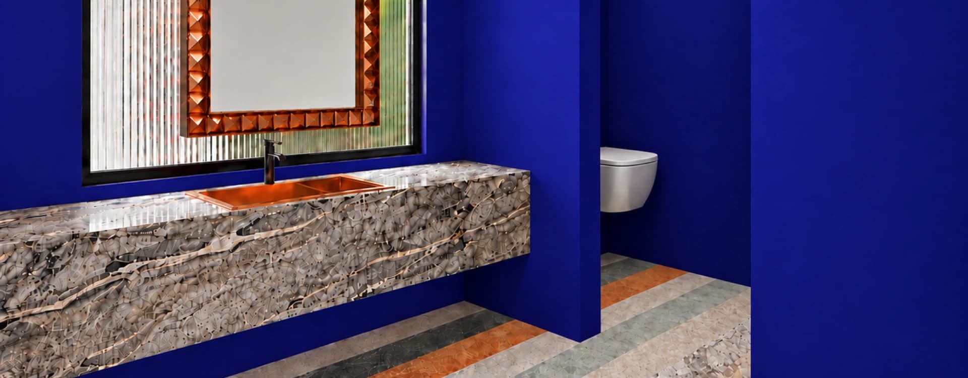 A cobalt-blue bathroom with a veined marble vanity, a copper sink and a carved copper-framed mirror, beside the toilet area