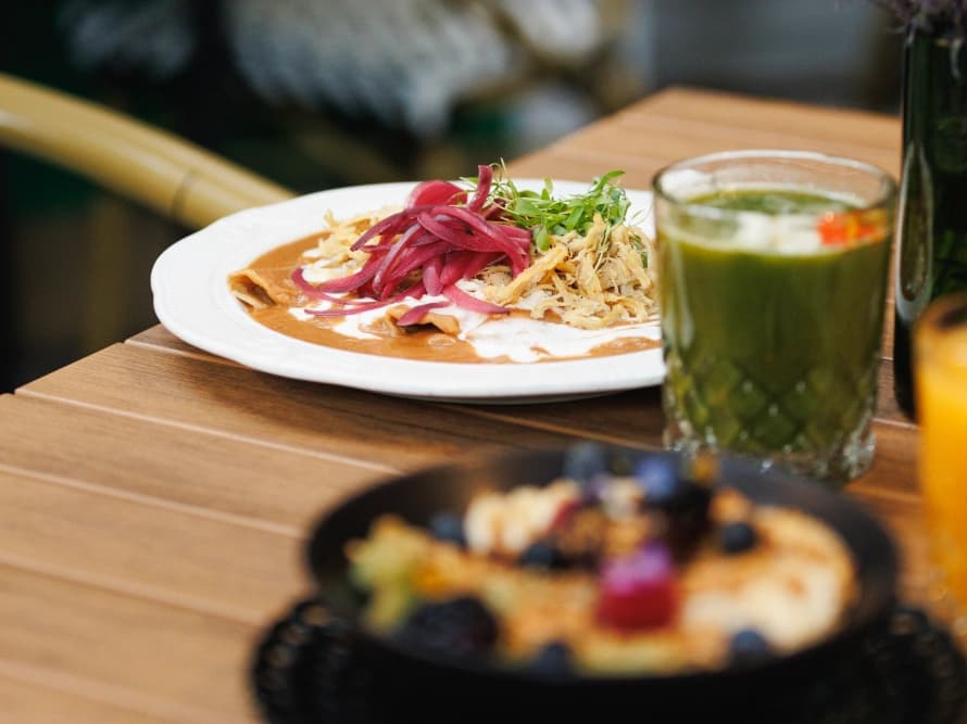 A marble table amid lush greenery, with a glass bottle of flowers and a plated sandwich