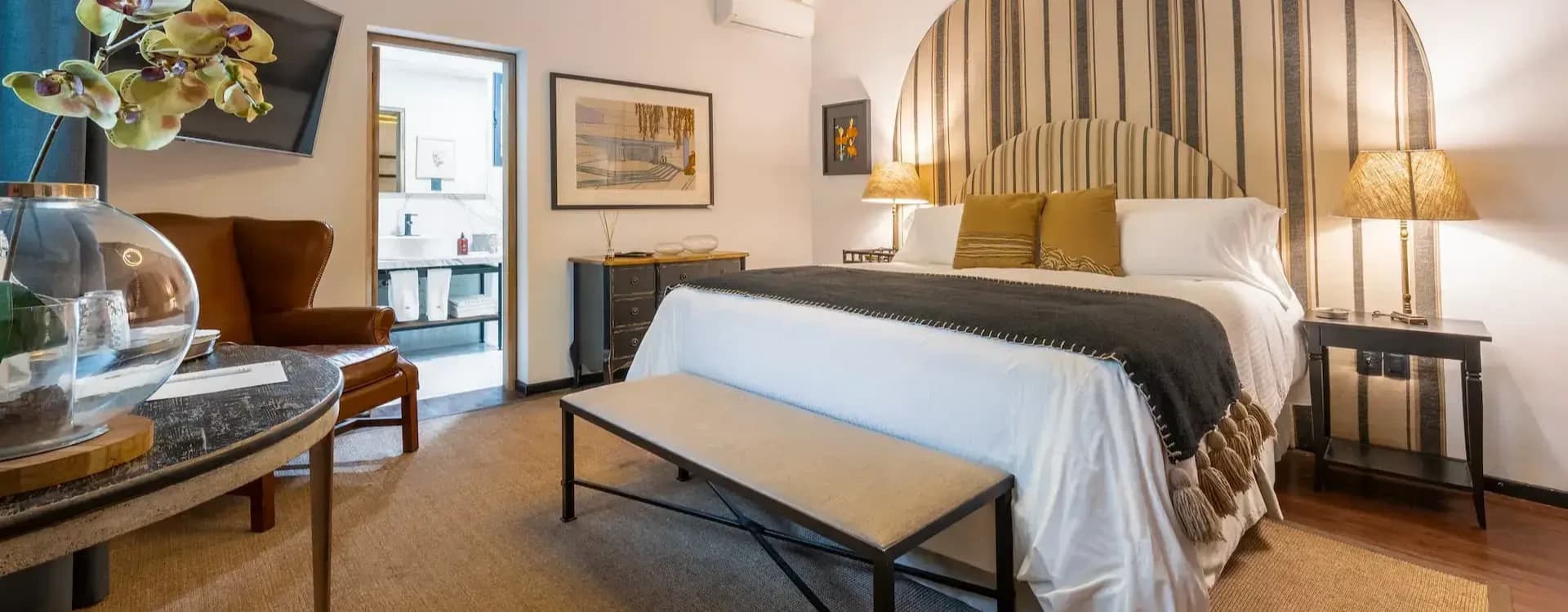 Room with a striped arched headboard, a wrought-iron bench and the bathroom beyond