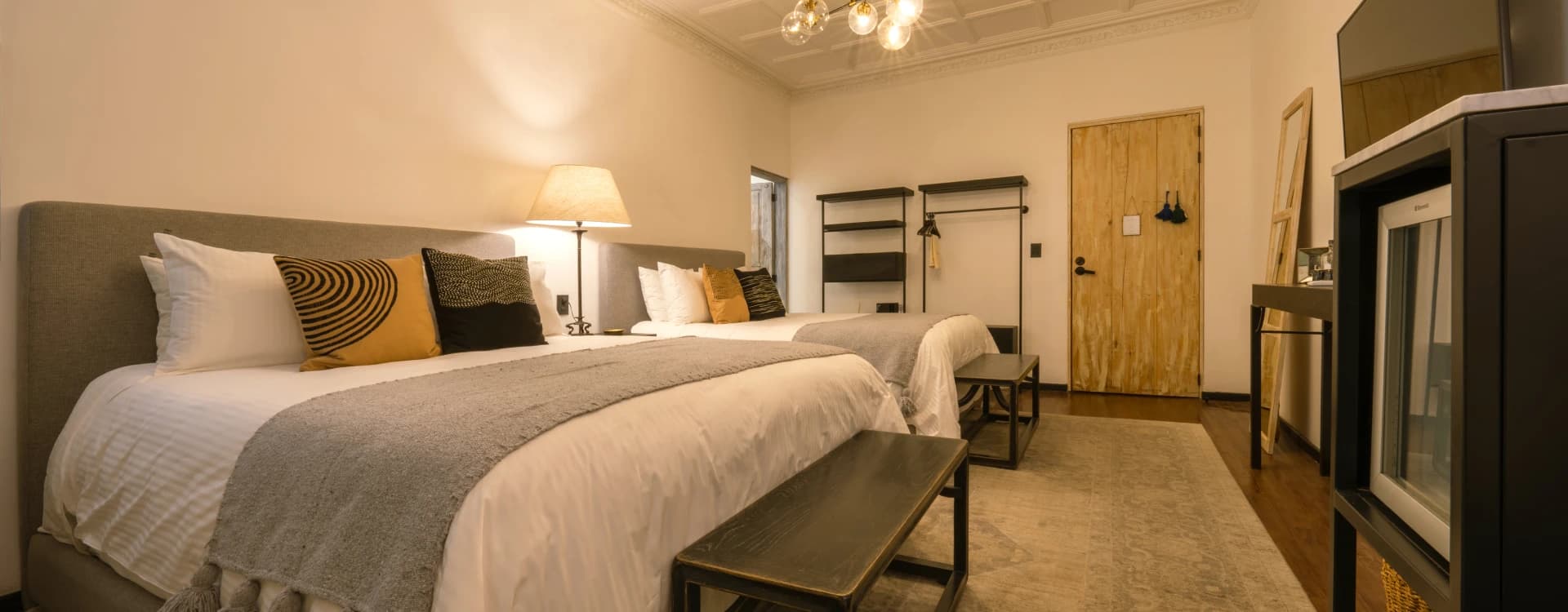 Two beds with grey headboards, a dark timber bench and a pale timber door beyond