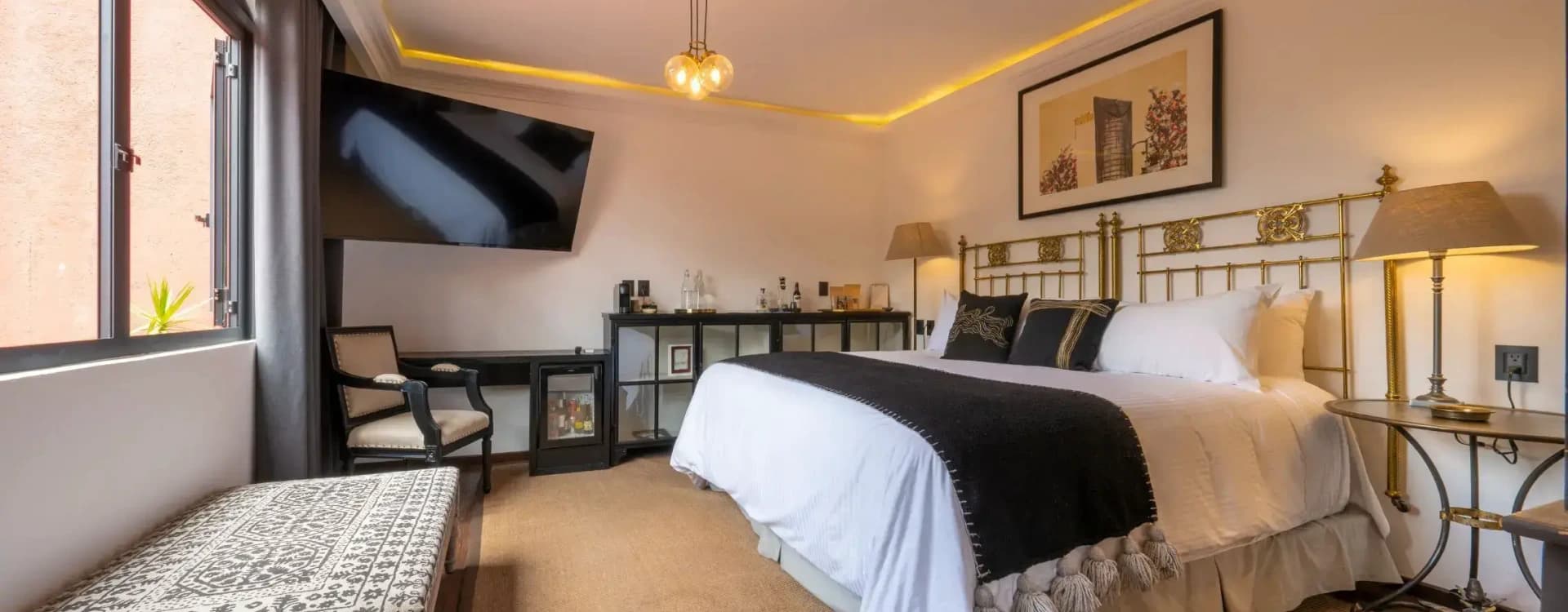 Bed with a brass headboard, a minibar, a desk and a screen under warm cove lighting