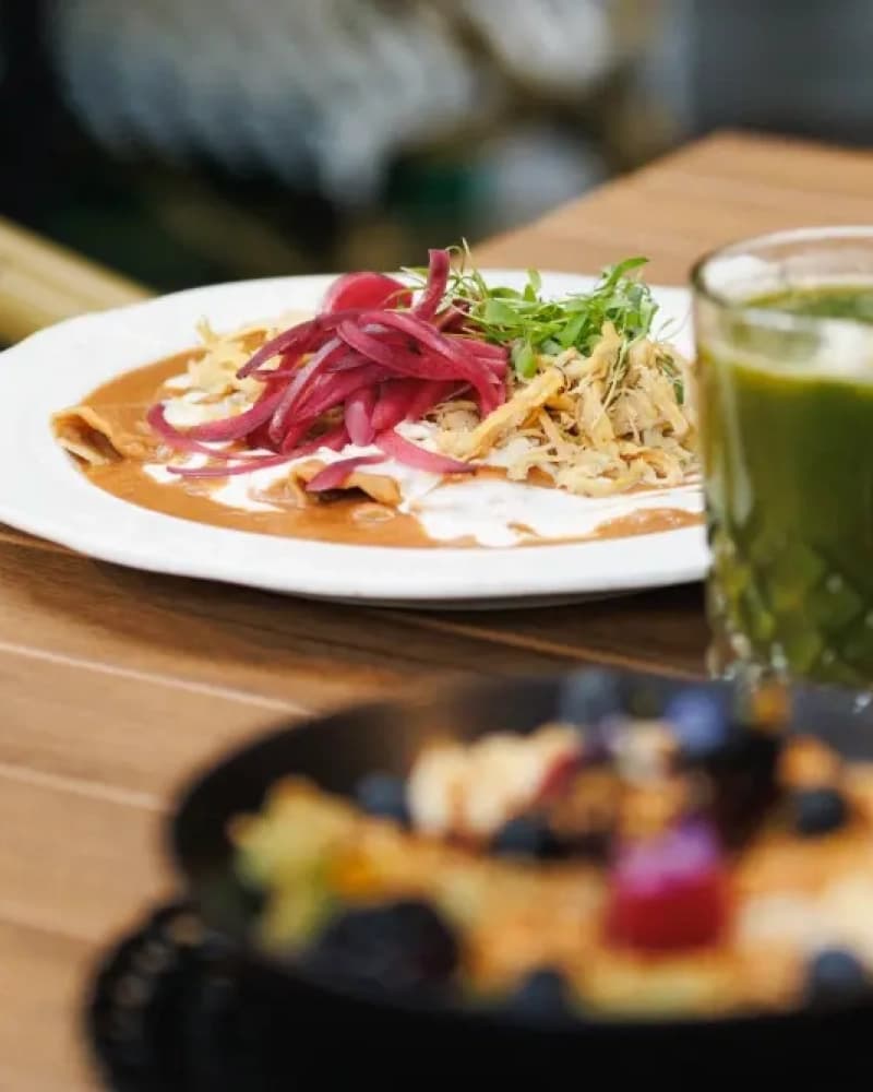 A plate of tortillas in sauce with red onion and cream, beside a green juice and a bowl of fruit, on a wooden table